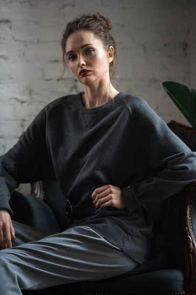 Atractiva morena mujer en gris ropa de moda sentado en el sillón y mirando a la cámara - foto de stock