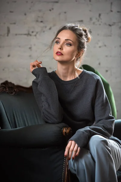 Atractiva mujer morena en jersey de moda sentada en sillón y mirando hacia otro lado - foto de stock