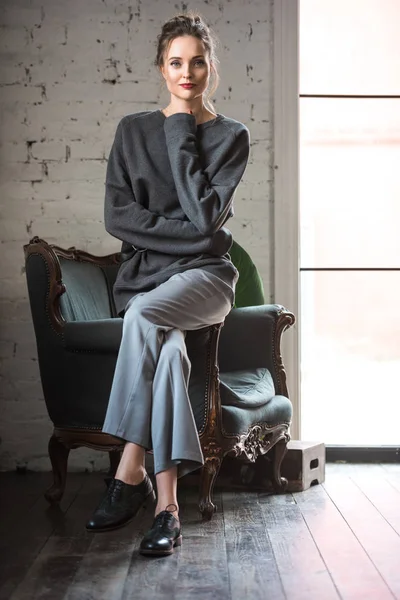 Vue pleine longueur de belle femme brune en tenue à la mode assise sur un fauteuil et souriant à la caméra — Photo de stock