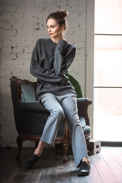 Full length view of beautiful brunette woman in fashionable outfit sitting on armchair and looking away — Stock Photo