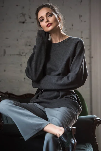 Beautiful tender girl in grey jumper and pants sitting on armchair and looking at camera — Stock Photo