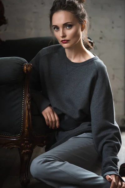 Hermosa mujer morena en ropa gris con estilo apoyado en el sillón y mirando a la cámara - foto de stock