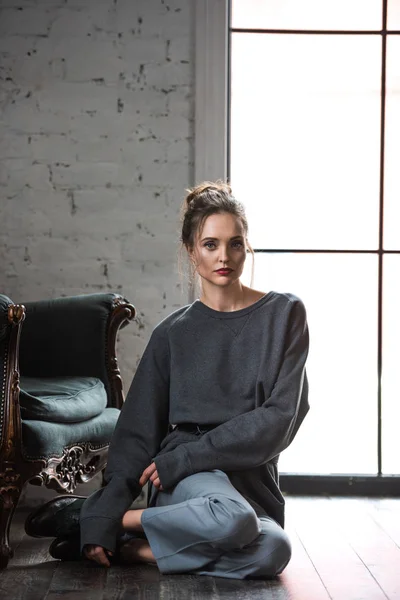 Beautiful brunette woman in fashionable outfit sitting on hardwood floor and looking at camera — Stock Photo