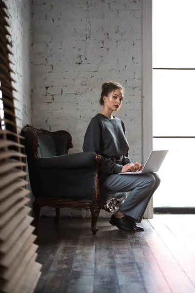 Vista completa de la hermosa mujer con estilo utilizando el ordenador portátil y mirando a la cámara mientras está sentado en el sillón - foto de stock