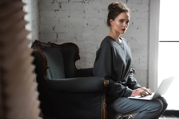 Hermosa mujer joven usando el ordenador portátil y mirando a la cámara mientras está sentado en el sillón - foto de stock