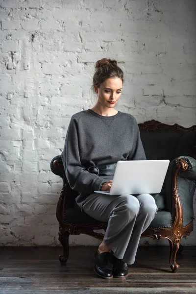 Beautiful brunette woman in stylish outfit using laptop while sitting on armchair — Stock Photo