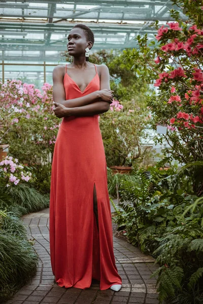 Fashionable african american model in red dress with crossed arms posing in garden with flowers — Stock Photo