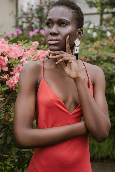 Chica afroamericana elegante con el pelo corto posando en vestido rojo en el jardín - foto de stock