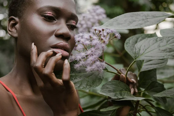 Stylish african american woman with glitter eyeshadow posing with flowers — Stock Photo
