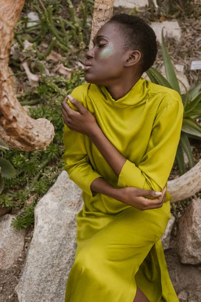 Tierna mujer afroamericana con los ojos cerrados y maquillaje brillo posando en jardín tropical - foto de stock