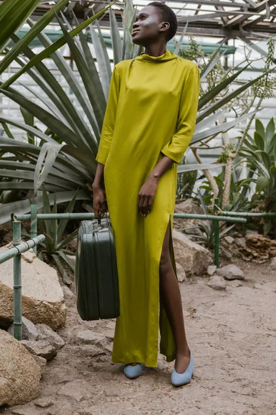 Mujer afroamericana con estilo en vestido amarillo posando con bolsa de viaje en invernadero tropical - foto de stock