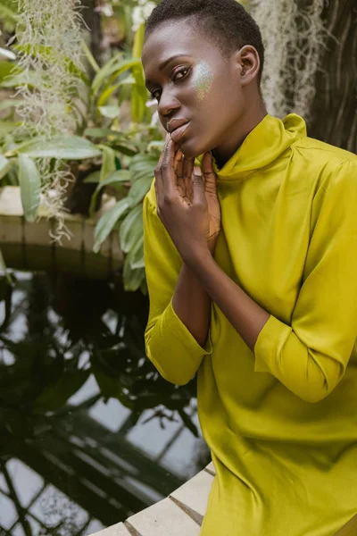 Tendre attrayant afro-américain femme avec paillettes maquillage posant près de la piscine dans le jardin tropical — Photo de stock