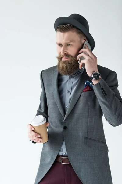 Elegante hombre de negocios barbudo sosteniendo una taza de papel y hablando por teléfono aislado sobre un fondo claro - foto de stock