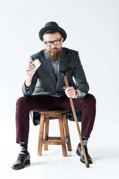 Elegante hombre de negocios barbudo sosteniendo dólares y caña mientras está sentado en el taburete - foto de stock