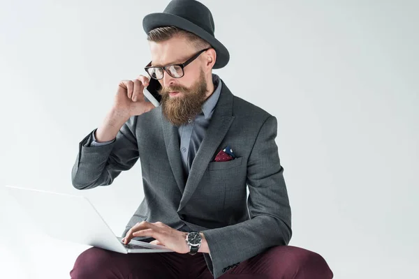 Stylish bearded businessman talking on smartphone and working on laptop isolated on light background — Stock Photo