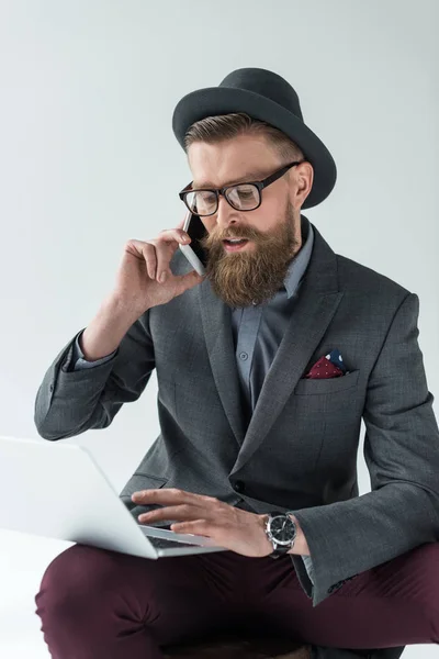 Geschäftsmann mit Vintage-Schnurrbart und Bart spricht auf Smartphone und arbeitet isoliert auf hellem Hintergrund am Laptop — Stockfoto