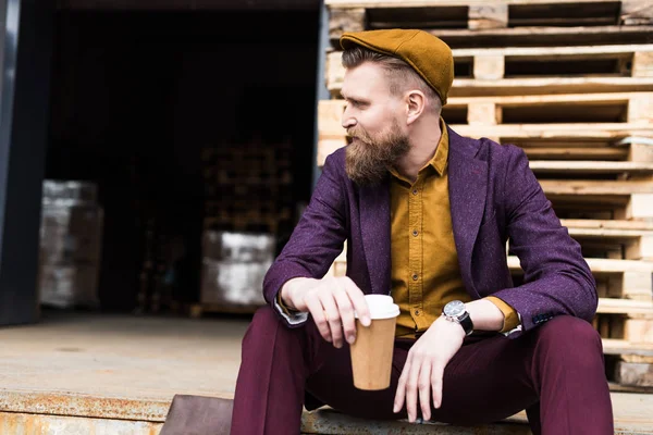 Elegante hombre de negocios barbudo sentado en la calle con taza de papel en las manos - foto de stock