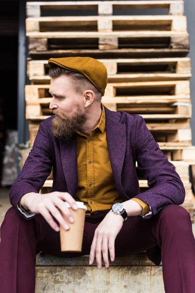 Empresario con bigote vintage y barba sentado en la calle con taza de papel en las manos - foto de stock