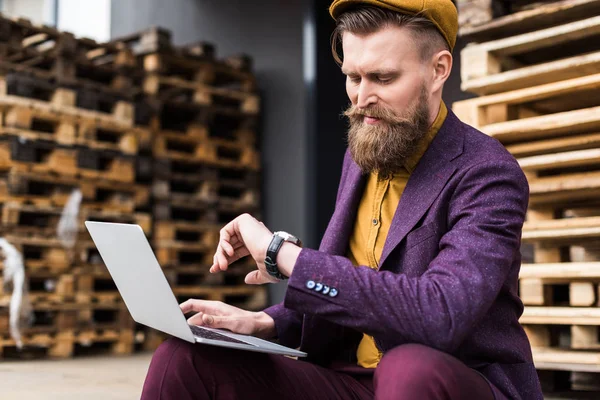 Stylish bearded businessman working on laptop on street and checking his watch — Stock Photo
