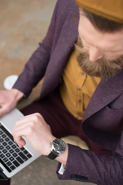 Empresario con bigote vintage y barba trabajando en el portátil y revisando su reloj - foto de stock