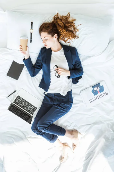 Vista dall'alto della donna d'affari in giacca e cravatta che tiene la tazza di caffè e dorme nel letto tra cartelle e documenti — Foto stock