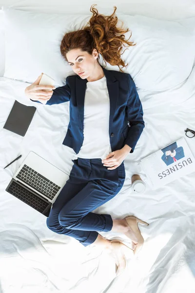 Von oben: Geschäftsfrau im Anzug macht Selfie im Bett zwischen Ordnern und Dokumenten — Stockfoto