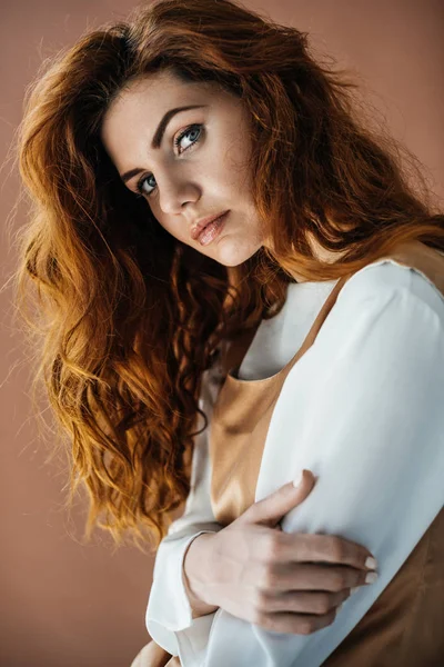 Donna con lunghi capelli rossi in abito beige guardando la fotocamera isolata su sfondo marrone — Foto stock