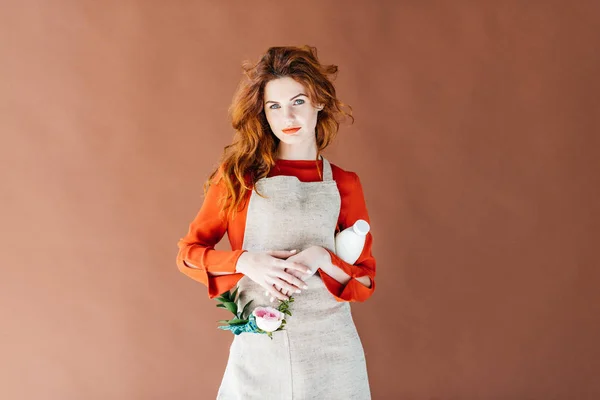 Hermosa joven en delantal sosteniendo botella de leche aislada sobre fondo marrón - foto de stock