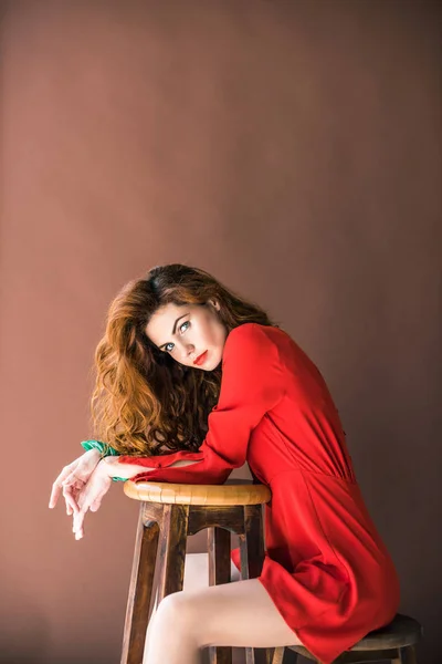 Woman with long red hair sitting on chair by stool isolated on brown background — Stock Photo