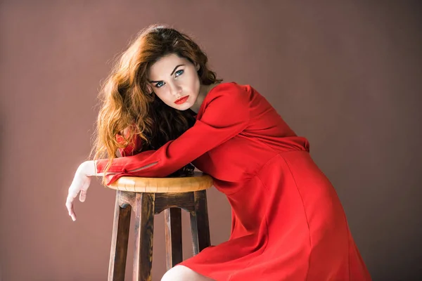 Attractive redhead woman posing by wooden stool isolated on brown background — Stock Photo