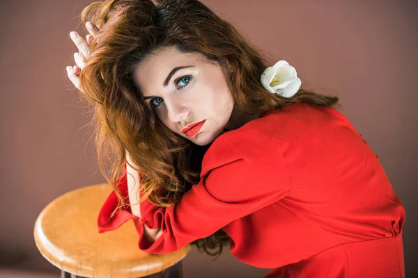 Mujer de pelo largo y rojo con flor de tulipán apoyada en taburete aislada sobre fondo marrón - foto de stock