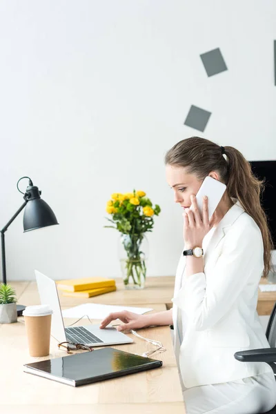 Vista lateral de la mujer de negocios en traje elegante hablando en el teléfono inteligente mientras se utiliza el ordenador portátil en el lugar de trabajo en la oficina - foto de stock