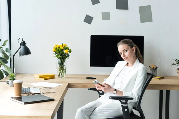 Businesswoman in stylish suit with smartphone at workplace with laptop in office — Stock Photo