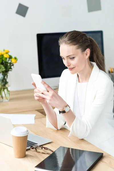 Усміхнена бізнес-леді в стильному костюмі, використовуючи смартфон на робочому місці в офісі — стокове фото