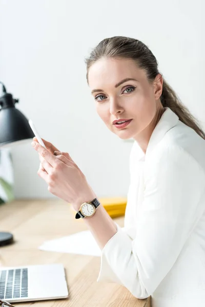 Side view of businesswoman in stylish suit with smartphone at workplace with laptop in office — Stock Photo
