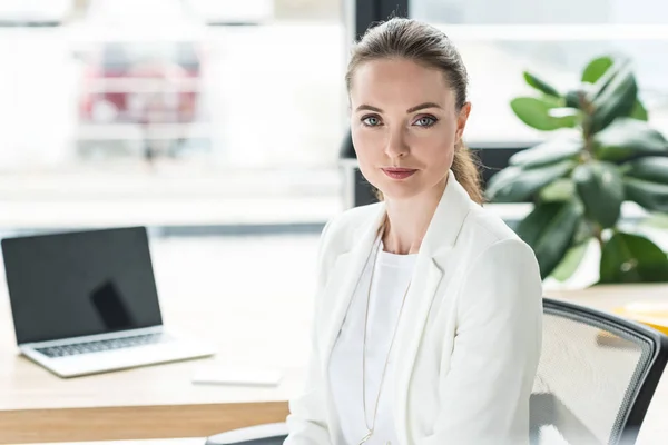 Portrait of smiling beautiful businesswoman in white suit at workplace with laptop in office — Stock Photo