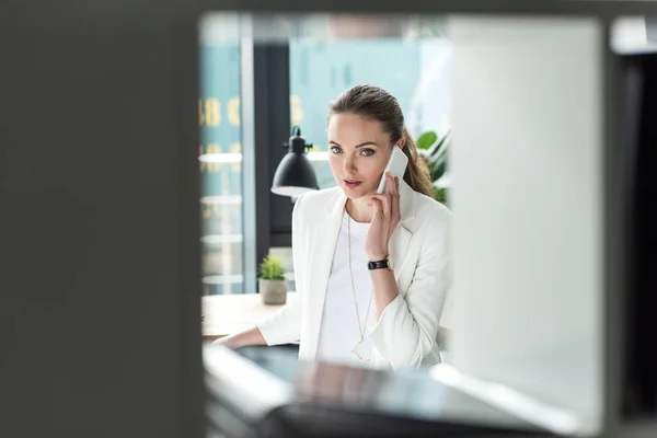 Selective focus of businesswoman in stylish suit talking on smartphone at workplace in office — Stock Photo