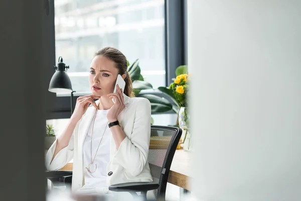 Selektiver Fokus einer Geschäftsfrau im schicken Anzug, die am Arbeitsplatz im Büro mit dem Smartphone spricht — Stockfoto