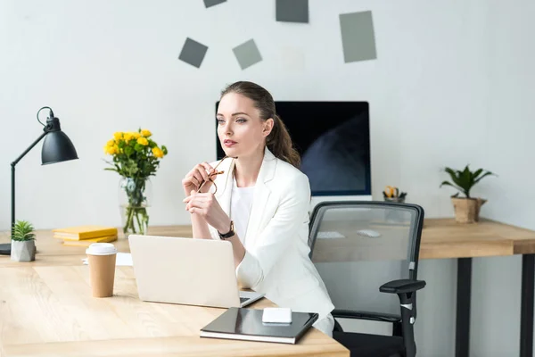 Belle femme d'affaires coûteuse en costume blanc regardant loin sur le lieu de travail avec ordinateur portable au bureau — Photo de stock