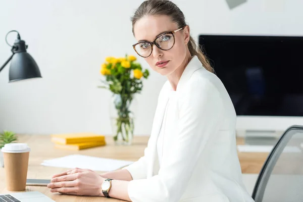 Vista lateral de hermosa mujer de negocios en traje blanco y gafas en el lugar de trabajo en la oficina - foto de stock