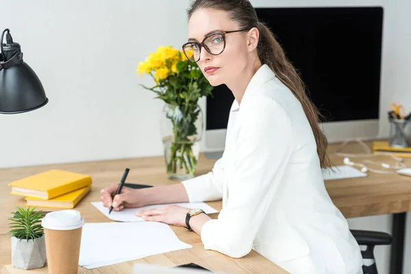 Donna d'affari in occhiali che guarda la fotocamera mentre fa scartoffie sul posto di lavoro in ufficio — Foto stock