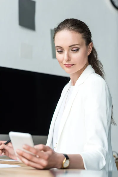 Selective focus of beautiful businesswoman using smartphone at workplace in office — Stock Photo