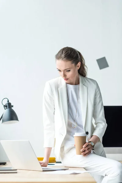 Retrato de empresária atraente com café para ir usando laptop no local de trabalho no escritório — Fotografia de Stock