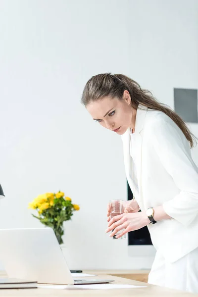 Donna d'affari focalizzata con vetro d'acqua che guarda lo schermo di computer portatile a posto di lavoro in ufficio — Foto stock