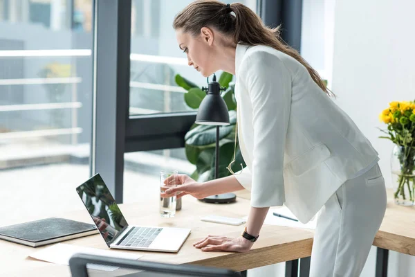 Visão lateral de empresária com vidro de água usando laptop no local de trabalho no escritório — Fotografia de Stock