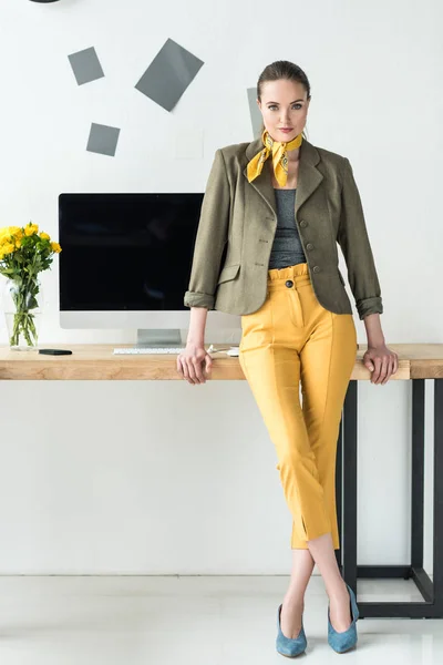 Hermosa mujer de negocios en ropa de moda apoyada en el lugar de trabajo con pantalla de computadora en la oficina - foto de stock