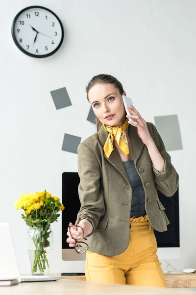 Hermosa mujer de negocios elegante con gafas y hablando por teléfono inteligente en la oficina - foto de stock