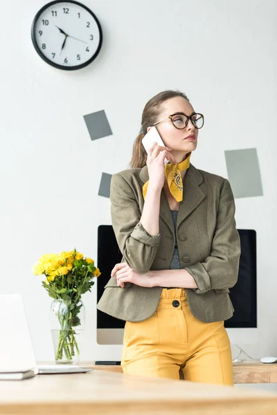 Attractive businesswoman in eyeglasses talking by smartphone and looking away in office — Stock Photo