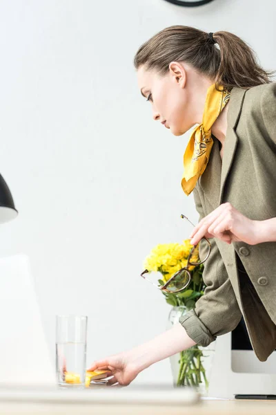 Side view of stylish businesswoman holding eyeglasses and reaching for glass of water in office — Stock Photo