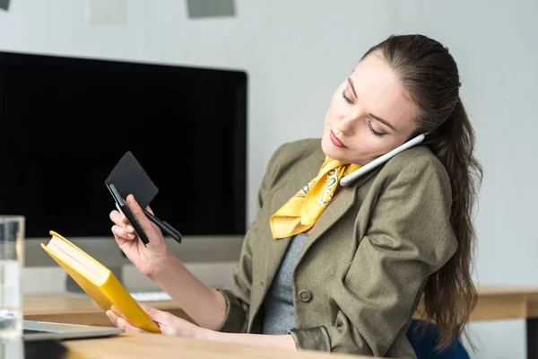 Geschäftsfrau spricht per Smartphone, während sie Buch und Taschenrechner am Arbeitsplatz in der Hand hält — Stockfoto